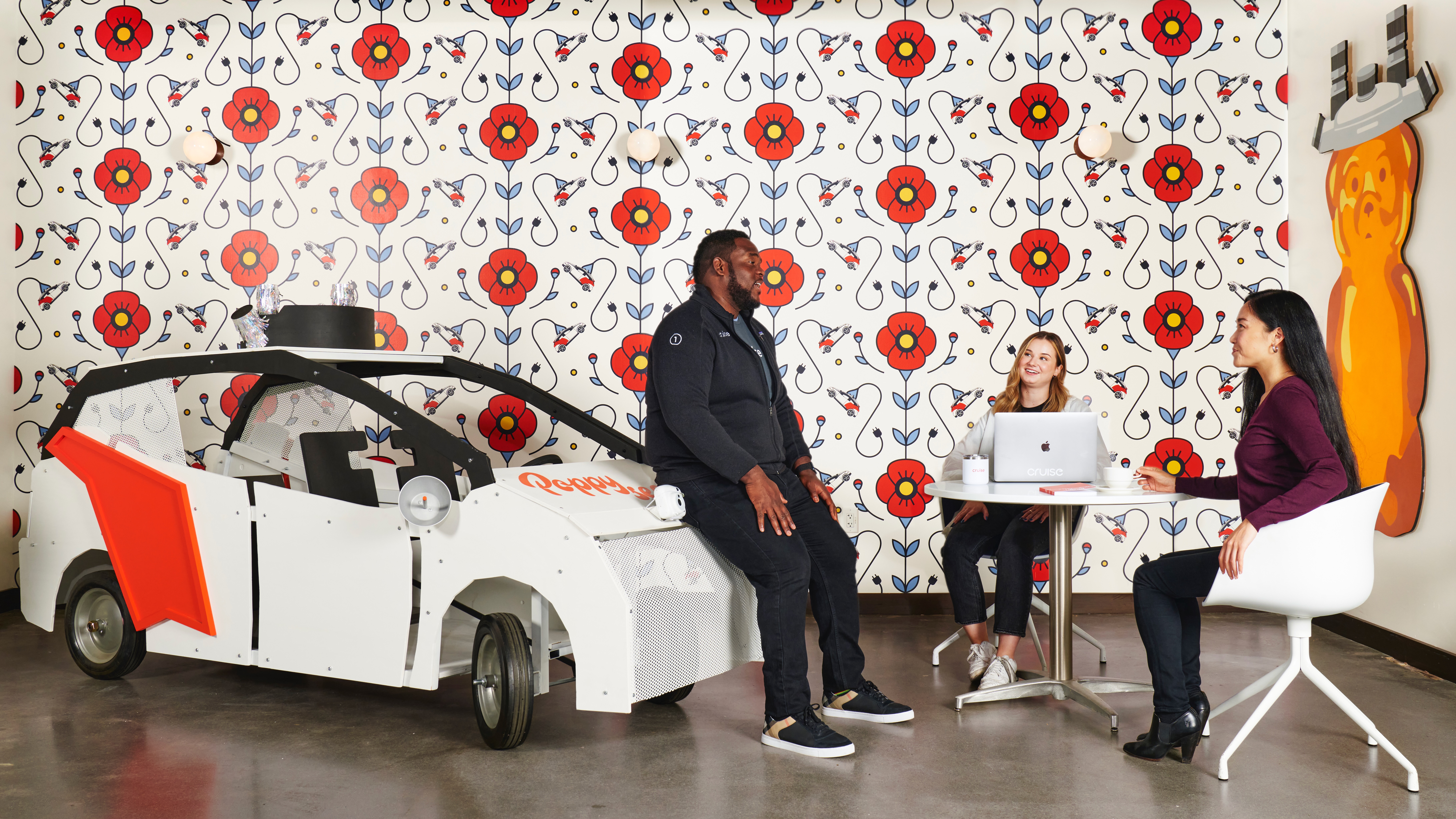 Three colleagues talk inside Poppy's Cafe in Cruise's HQ, one is sitting on the front of a Cruise driverless vehicle model