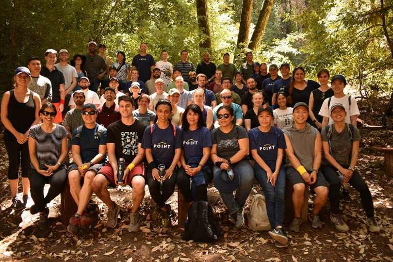 A large group of Point employees pose together for a group photo in the forest during a team outing.