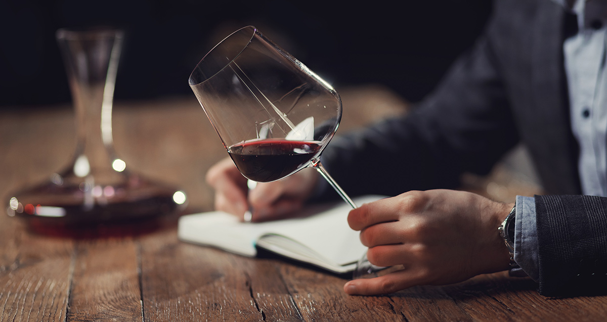 Male sommelier holding a glass of red wine writing tasting notes in a journal