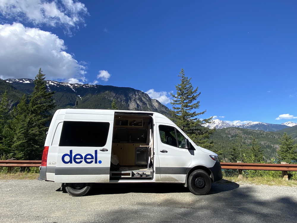 A white van with the blue Deel logo parked in front of a mountain view.