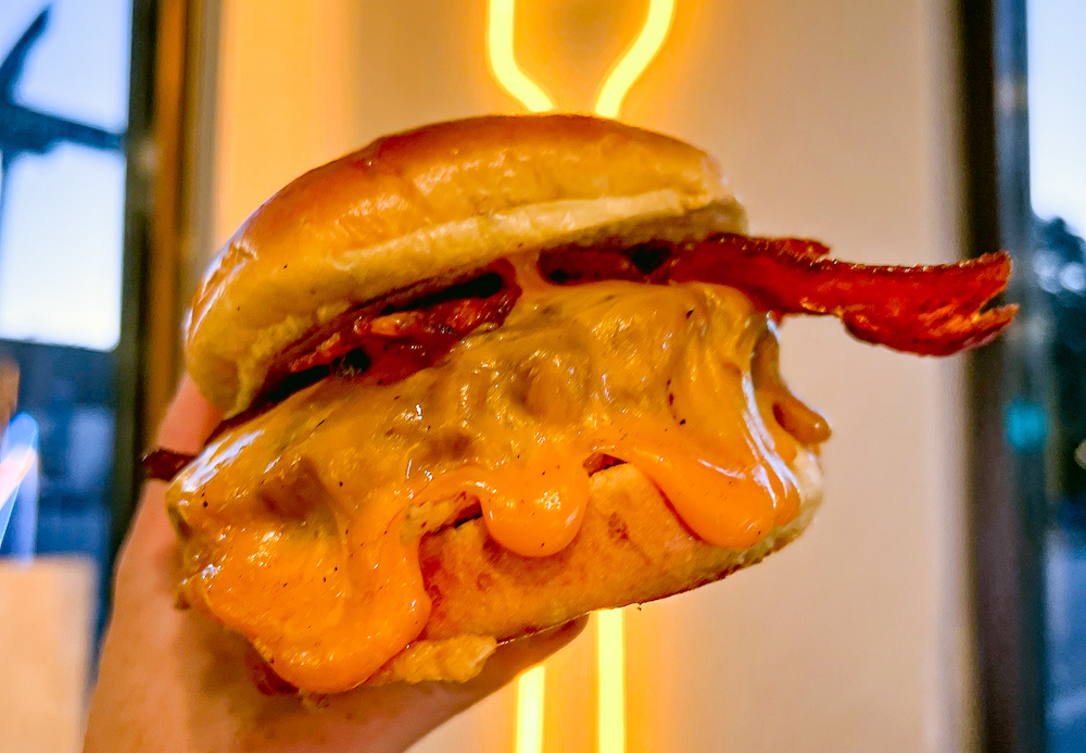 a gooey cheeseburger held in front of a neon sign