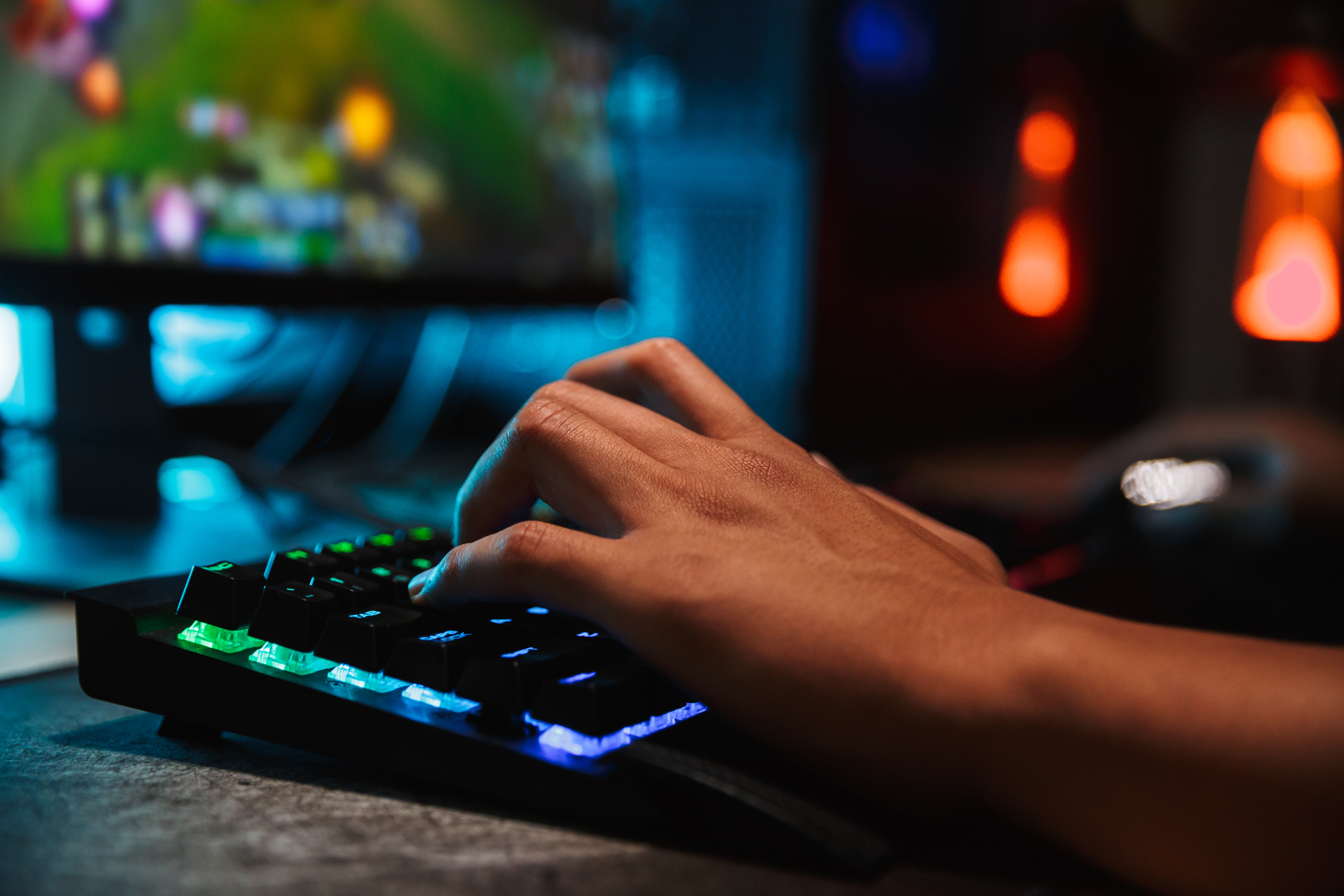 Image of a person hands on an illuminated keyboard in front of an out-of-focus computer displaying a video game