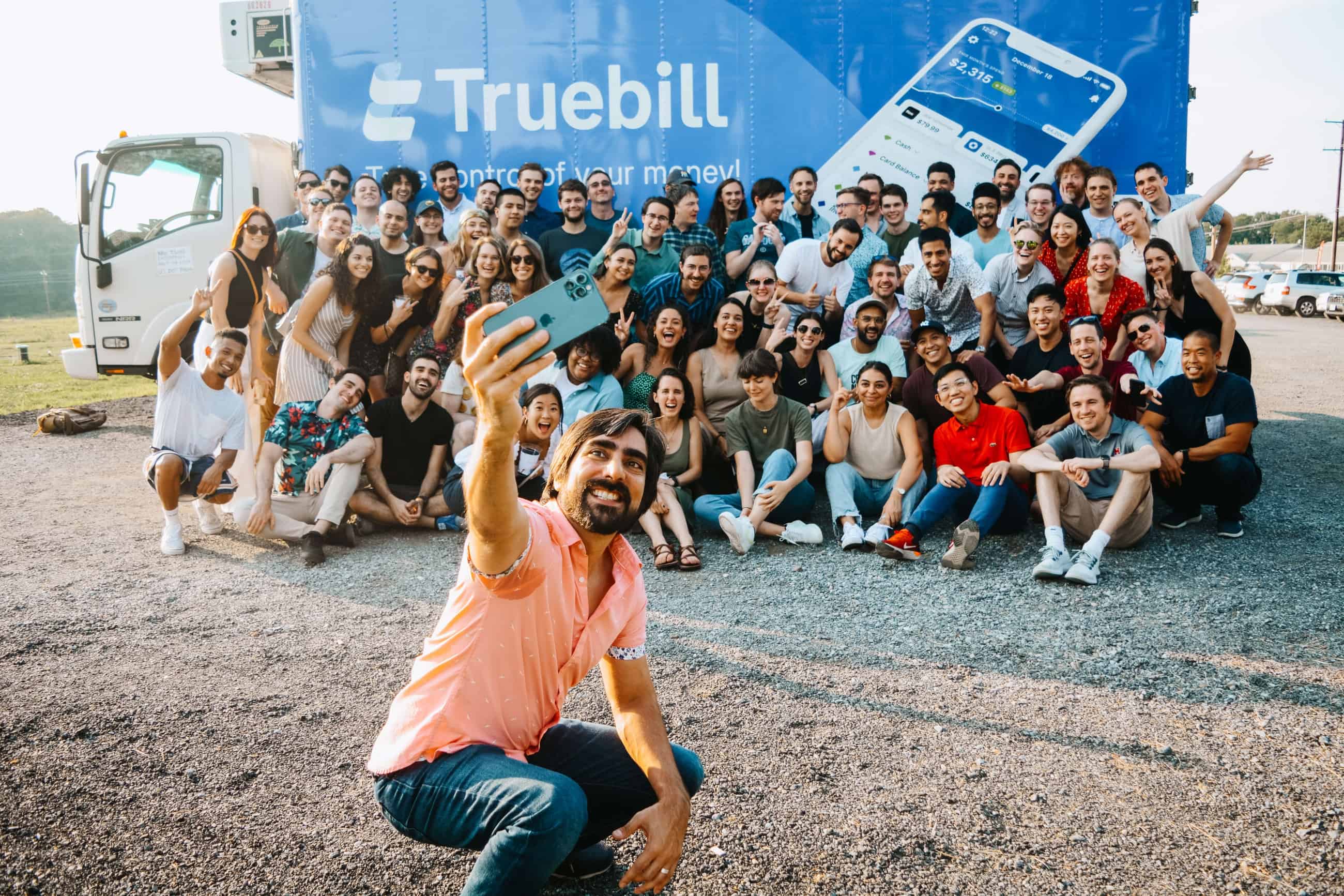 A photo of a Truebill employee taking a selfie with a large number of people in front of a truck bearing the Truebill logo.